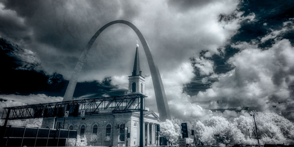 Elevation of Timeless Symmetry beckons with a spellbinding allure, weaving a tale where history and modernity coalesce in a singular, monochromatic narrative. Awash in spectral tones, this infrared photograph transports viewers to a realm where the ethereal and tangible intersect, sculpting a dreamscape that defies the boundaries of time.  At the heart of this visual symphony stands a dignified historical building, its steeple piercing the heavens with an air of solemn reverence. The architectural grace of this heritage structure whispers secrets of bygone eras, grounding the viewer in a tradition that feels both eternal and fleeting. Towering behind it, an arching monolith of contemporary design rises, a guardian of progress and innovation. This juxtaposition of old and new creates a dance of lines and forms, harmonizing the essence of past and future.  The sky above, alive with brooding, textured clouds, commands the scene with a dramatic flair. Voluminous and ever-shifting, these clouds cast ever-changing shadows that breathe life into the monochrome palette, adding layers of mystery and depth. The absence of color turns the familiar into the fantastical, endowing the scenery with a surreal, almost otherworldly quality.  Through the lens of infrared light, the photograph transcends its physical constraints, becoming an enigmatic portal where tradition and modernity are not opposing forces but complementary echoes of an unfolding myth. Elevation of Timeless Symmetry invites its audience to linger, to ponder the seamless blend of epochs, and to lose themselves in a contemplation of what was, what is, and what might yet be.