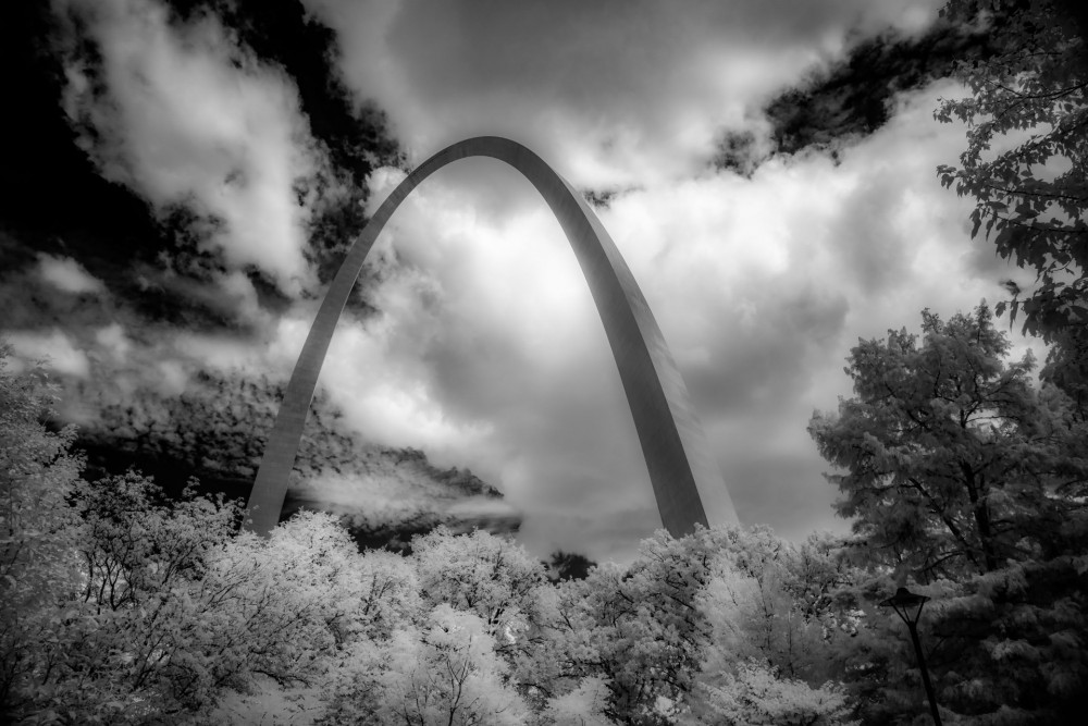 This evocative monochrome photograph, titled "Infinite Curves in Saint Louis," captures the majestic Gateway Arch in a way that feels both timeless and powerful. The arch ascends magnificently into a textured sky, its sleek, curved structure standing in stark contrast against the dramatic backdrop of swirling clouds. This infrared artwork transforms the scene, lending an ethereal quality to the composition, as the natural beauty of the surrounding trees frames the bottom of the image and introduces depth and context.  The choice of a black-and-white palette amplifies the artwork's dramatic impact, stripping the scene down to its most essential elements and allowing each detail to shine with clarity. The viewer is drawn into a moment that transcends time, where the natural world and human achievement blend harmoniously. This photograph does more than capture a landmark; it elevates it into a symbol of aspiration and unity, inviting the audience to reflect on the infinite possibilities that lie within the seamless blend of nature and ingenuity.