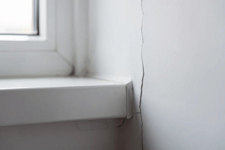 A close-up view shows a small hairline crack along a white interior wall near a window frame, indicating potential house settling issues. These minor cracks can be part of the normal settling process, but may also suggest underlying problems with the home's foundation or the soil conditions beneath.