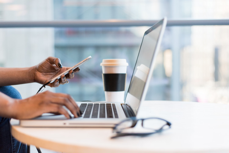 close up of side view of a laptop with a woman's hands on it, next to it is a paper coffee cup and a computer mouse.